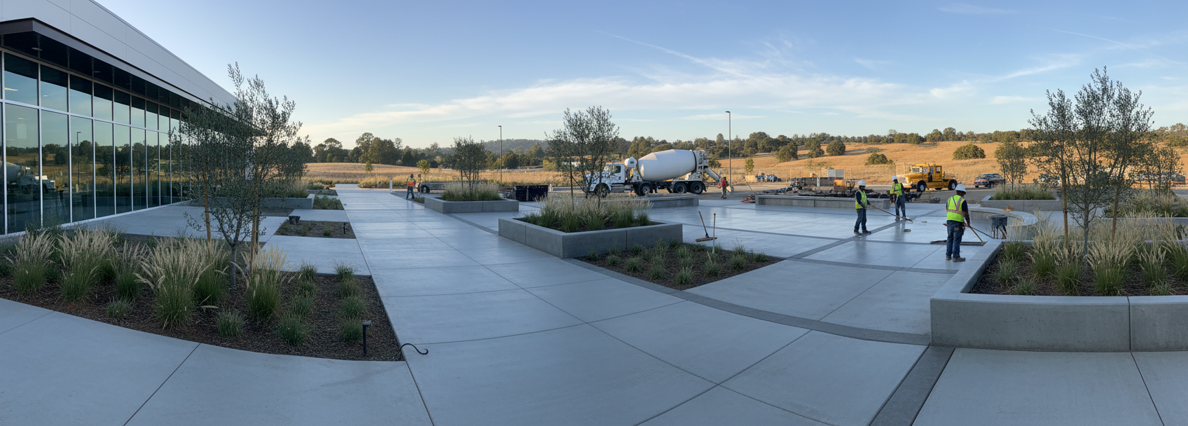Concrete construction site with workers, a cement truck, and new gray walkways next to a modern building, showing commercial